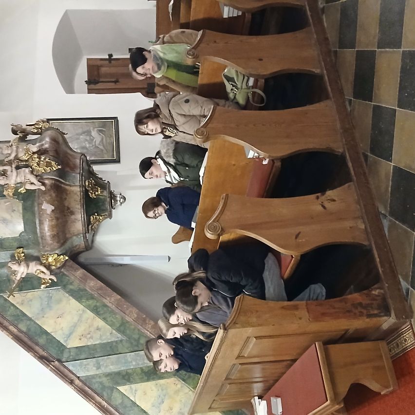 Eine Gruppe von Menschen sitzt in hölzernen Kirchenbänken. Ein kunstvoller Altar und ein gerahmtes Bild schmücken die Wand.