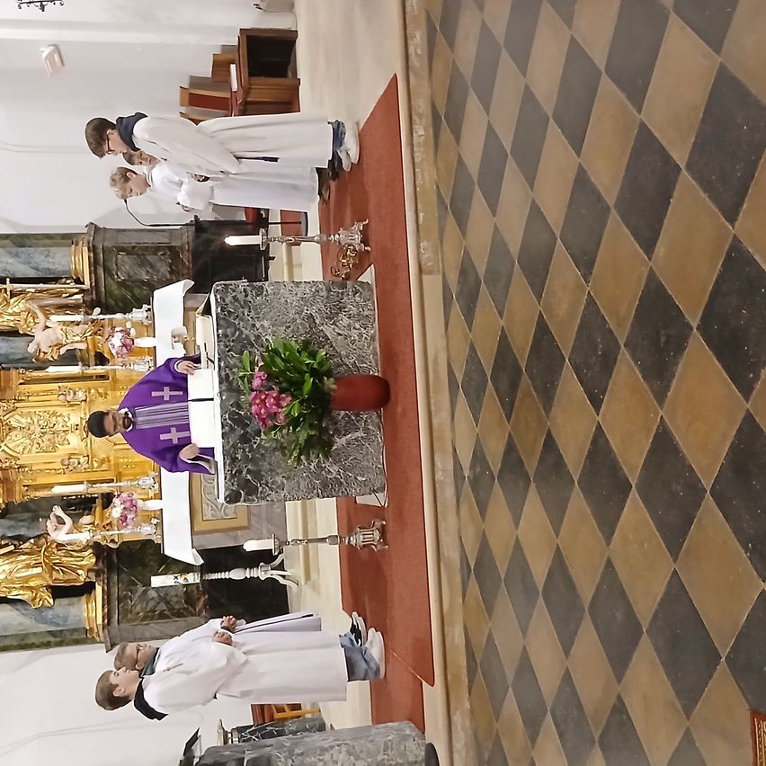 Ein Priester steht an einer Kanzel und liest aus einem Buch. Zwei junge Jungen, wahrscheinlich Altardiener, stehen hinter ihm. Der Altar ist mit Kerzen und Blumen geschmückt. Die Szene spielt sich in einer Kirche mit gemustertem Boden und einer goldenen Statue ab.