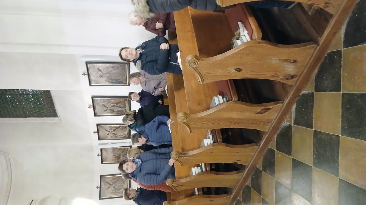 Eine Gruppe von Menschen steht in einer Kirche und schaut nach oben. Sie tragen Winterjacken. Die Kirche hat Holzbänke und mehrere Gemälde an der Wand.