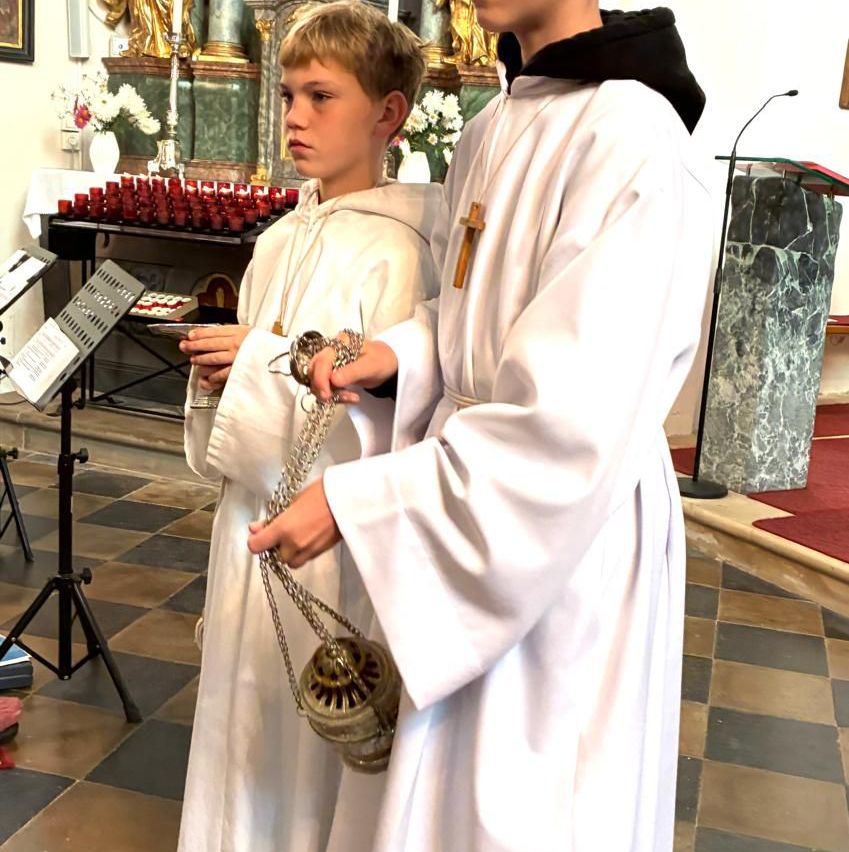 Two boys dressed in white robes stand in a church, one holding a ceremonial chain. They are in front of an altar with candles and a statue in the background.