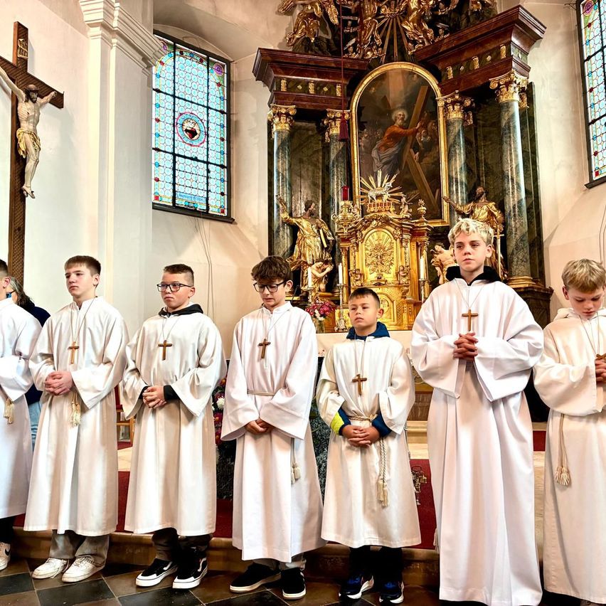 A group of boys dressed in white robes and holding crosses are standing in front of an altar. Behind them is a wall with a cross and a stained glass window.