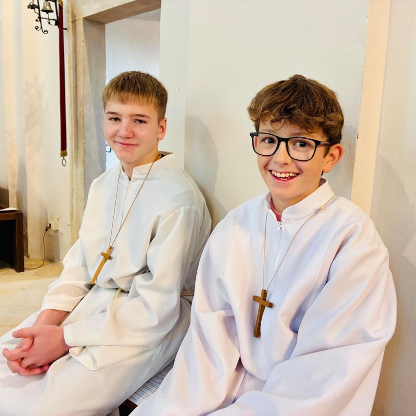 Two boys wearing white cassocks with cross necklaces sit next to each other. They appear to be smiling for a photo inside a church.
