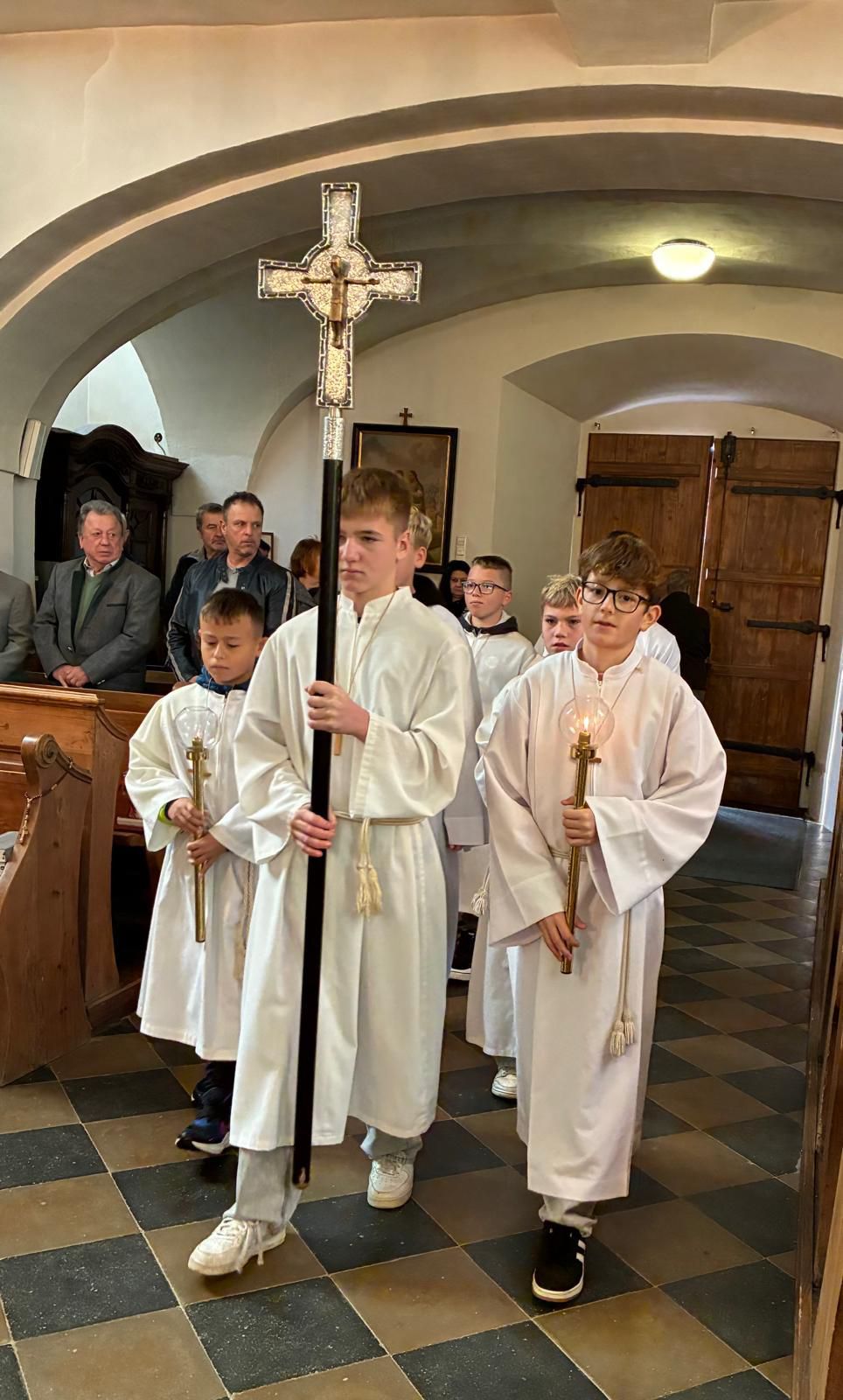 A group of young boys in white robes hold candles and a cross inside a church, with people standing behind them.