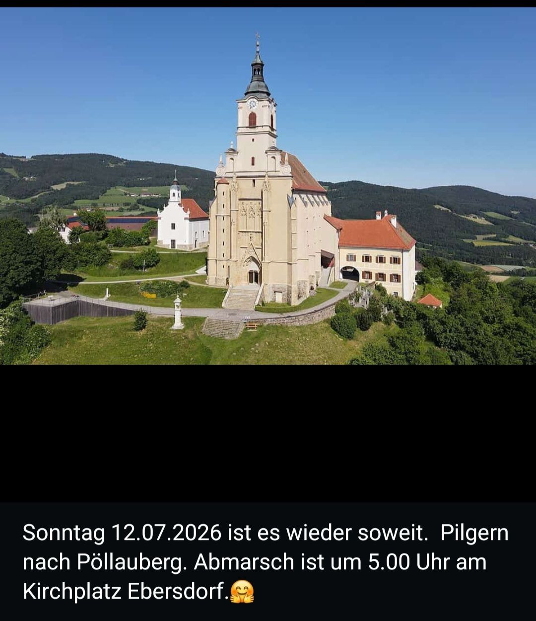 Aerial view of a church with a tall tower, surrounded by greenery. A smaller white building is visible in the background.
