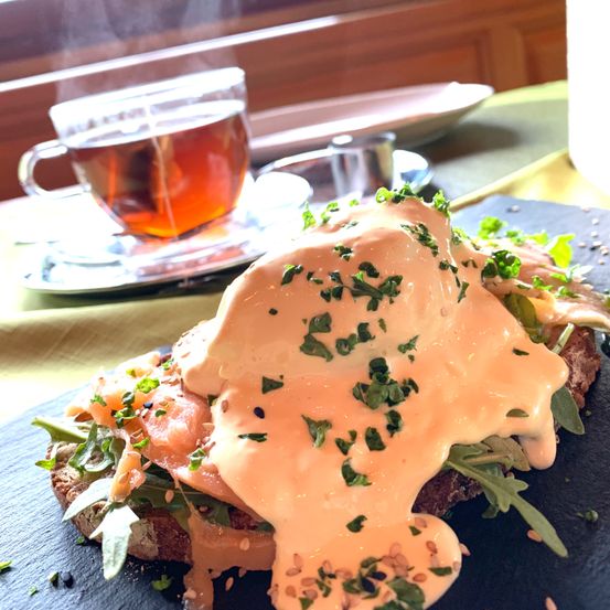 Ein Nahaufnahme eines Frühstücksgerichts mit pochiertem Ei, Lachs und Rucola auf einem dunklen Teller, begleitet von einer Tasse Tee auf einer Untertasse.