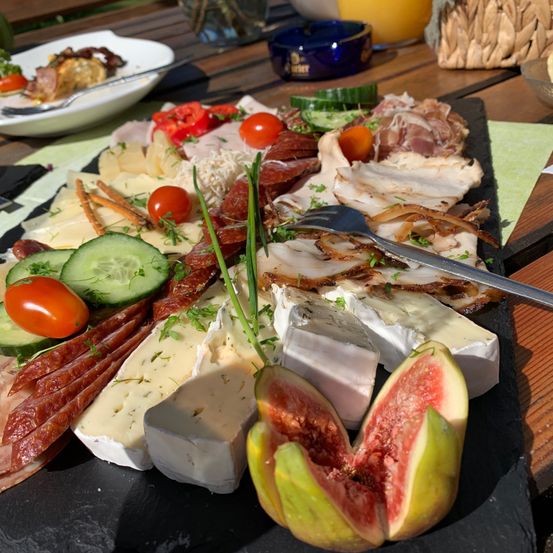 Eine Charcuterie-Platte mit verschiedenen Käsesorten, geschnittenem Fleisch, Tomaten, Gurken und Feigen, auf einem schwarzen Schiefer mit einer Gabel.