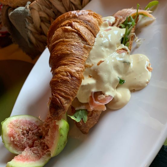 Ein Croissant-Sandwich mit Räucherlachs, Sahnesauce und Rucola auf einem weißen Teller, begleitet von einer geschnittenen Feige.