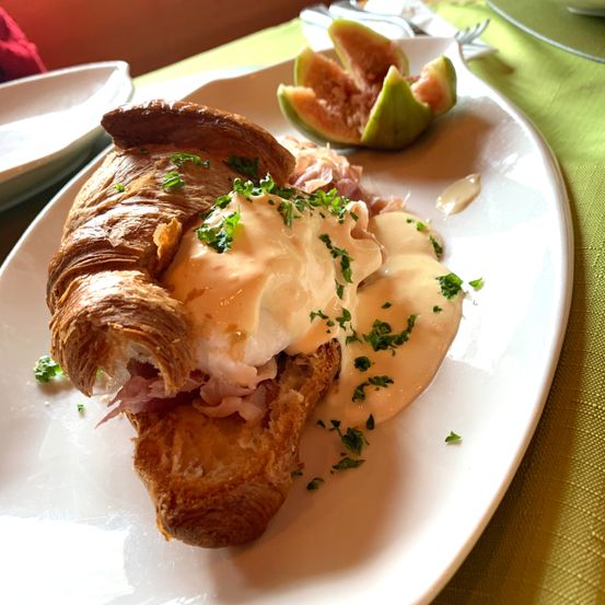 Ein Croissant-Sandwich mit pochiertem Ei, Schinken und Hollandaise-Sauce auf einem weißen Teller mit grüner Tischdecke.