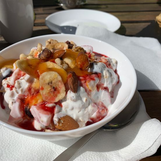 Eine weiße Schüssel mit einer cremigen Joghurt-Frucht-Salat, gekrönt mit Nüssen und einem Sirup-Strudel, auf einem weißen Servietten auf einem Holztisch.