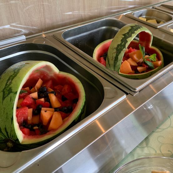 Zwei Wassermelonen in Herzform geschnitzt, gefüllt mit einer Mischung aus Wassermelonen, Cantaloupe, Blaubeeren und Minzblättern, auf einem Edelstahl-Buffetttablett.