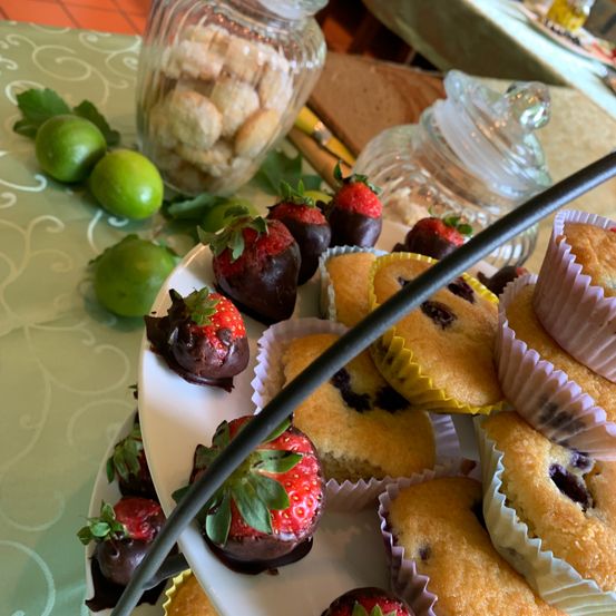 Ein weißer Teller mit verschiedenen Desserts, darunter schokoladenüberzogene Erdbeeren und Blaubeermuffins, auf einem Tisch mit Limetten und einem Glas mit Keksen im Hintergrund.