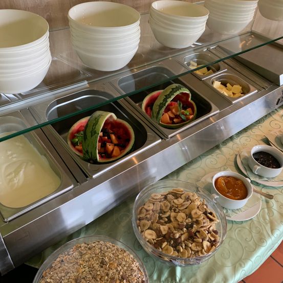 Ein Buffettisch mit verschiedenen Lebensmitteln, darunter eine Wassermelone und Obstsalat, Joghurt und eine Mischung aus Nüssen und Müsli.
