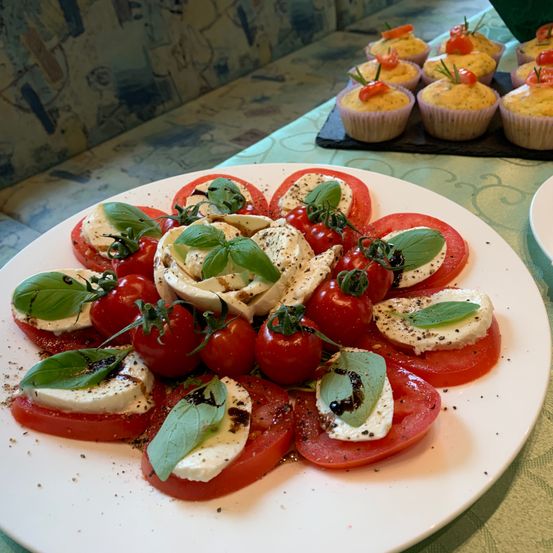 Ein weißer Teller mit einem Caprese-Salat aus geschnittenen Tomaten, Mozzarella, Basilikumblättern und Kirschtomaten, mit Cupcakes im Hintergrund.