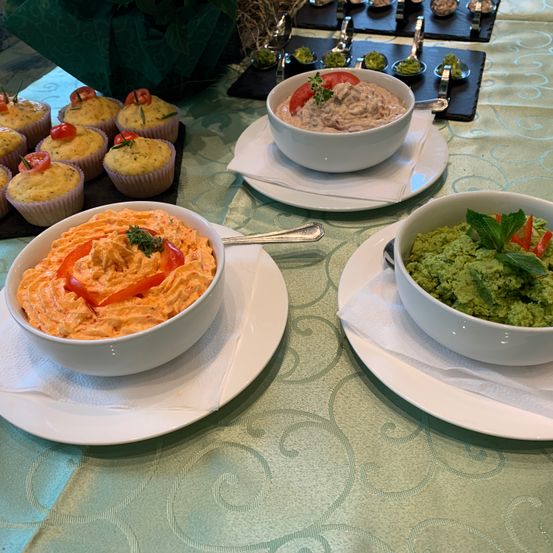 Ein Tisch mit verschiedenen Gerichten, darunter eine Schüssel mit oranger Dip, eine Schüssel mit grüner Dip und Cupcakes mit Tomaten obenauf.
