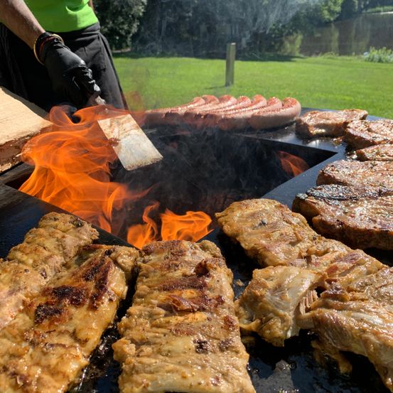 Eine Person grillt verschiedene Fleischsorten auf einem Grill mit Flammen, einschließlich Würste und Steaks, in einer Outdoor-Umgebung.