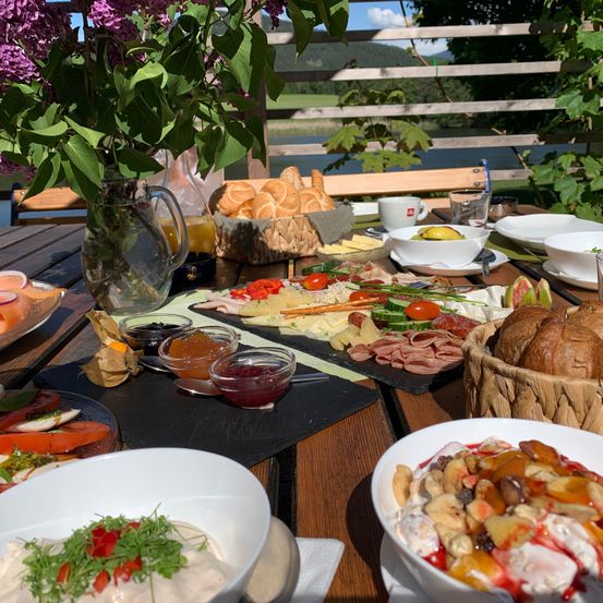 Ein Holztisch mit einer Auswahl an Speisen wie Brot, Käse, Fleisch und Salaten, mit einer Vase mit lila Blumen und einer malerischen Aussicht im Hintergrund.