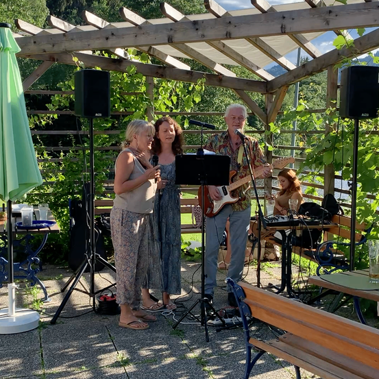Drei Personen treten auf einer Bühne unter einer hölzernen Pergola auf, einer spielt Gitarre und singt, während die anderen beiden Mikrofone halten.