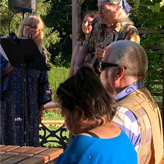 Eine Gruppe von Menschen führt eine musikalische Darbietung in einem Garten durch, wobei eine Frau in ein Mikrofon singt und ein Mann Gitarre spielt.