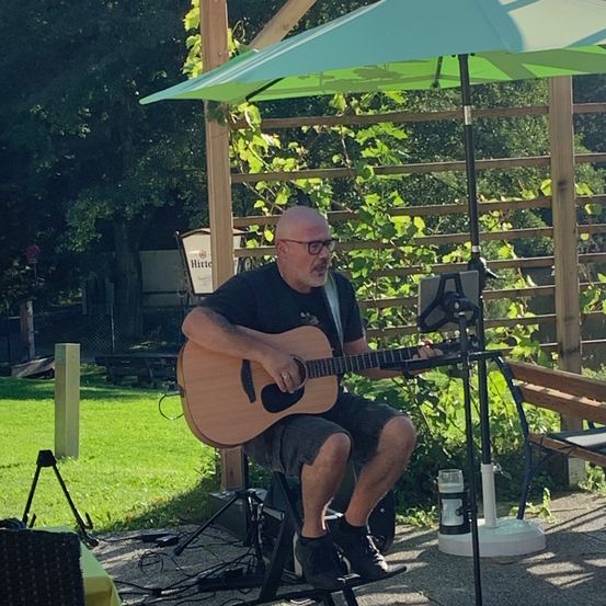 Ein Mann mit Brille und schwarzem Shirt spielt Gitarre unter einem grünen Schirm in einem Garten.