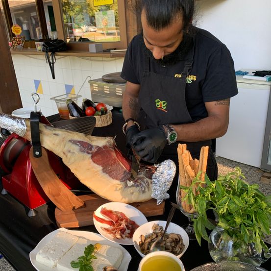 Ein Mann in einem schwarzen Apron und Handschuhen schneidet ein großes Stück Fleisch auf einem Holzbrett in einer Küche.