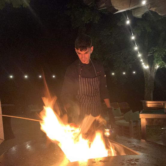 Ein Mann in einer Kochschürze kocht bei Nacht über einem offenen Feuer, mit sichtbaren Flammen und einem Stock.