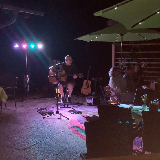 Ein Mann spielt Gitarre auf einer Bühne bei Nacht, beleuchtet von bunten Lichtern. Ein Publikum sitzt unter grünen Schirmen.