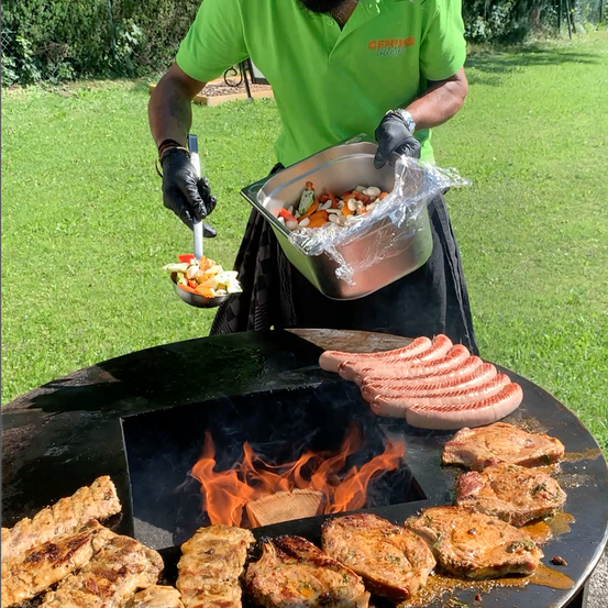 Eine Person in einem grünen Shirt grillt Fleisch und Gemüse auf einem großen Grill im Freien. Die Person hält einen Schöpflöffel mit Gemüse und eine Schale mit Plastikfolie.