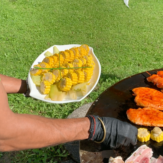 Eine Person hält einen Teller mit Maiskolben und einen weiteren Teller mit gegrilltem Hähnchen auf einem Grill.
