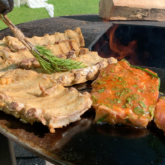 Ein Nahaufnahme von gegrillten Rippchen und einem Steak mit Kräutern auf einem Grill, mit einem Feuer im Hintergrund.