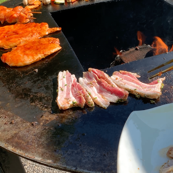 Ein Grill mit verschiedenen Fleischsorten, darunter Hühnchen und Speck, im Hintergrund brennt ein Feuer.