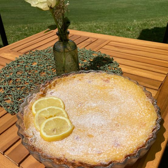 Eine Zitronentarte mit Puderzucker obenauf, auf einem Holztisch mit grünem Tuch und einer Vase mit einer weißen Blume.