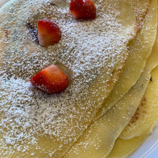 Ein Stapel von drei goldenen Crêpes, bestreut mit Puderzucker und geschnittenen Erdbeeren auf einem weißen Teller.