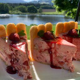 Ein Stück Erdbeer-Käsekuchen mit roter Sirup und frischen Erdbeeren auf einem weißen Teller, mit einem See und Bergen im Hintergrund.