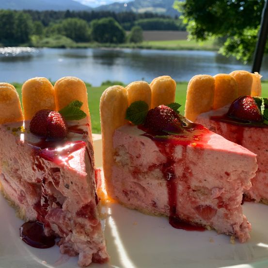Ein Stück Erdbeer-Käsekuchen mit roter Sirup und frischen Erdbeeren auf einem weißen Teller, mit einem See und Bergen im Hintergrund.
