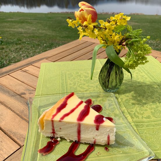 Ein Stück Käsekuchen mit roter Beerensauce auf einem Glasteller, begleitet von einer Vase mit gelben Blumen auf einem Holztisch mit einer malerischen Aussicht im Hintergrund.