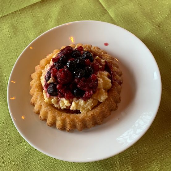 Eine kleine Tart mit knusprigem Boden, gefüllt mit cremigem Pudding und belegt mit einer Mischung aus roten und blauen Beeren auf einem weißen Teller.