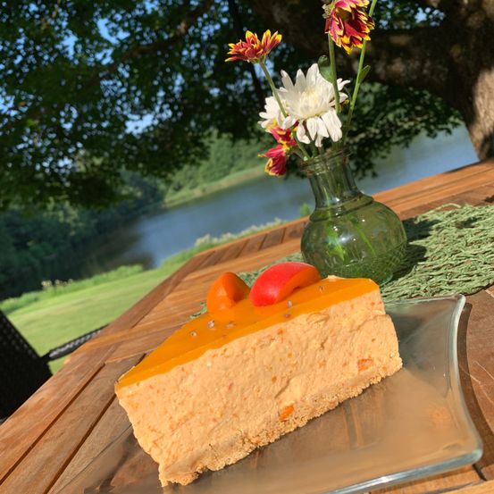 Ein Stück Karottenkuchen mit einem Pfirsich und einer Karotte darauf, auf einem Glasteller auf einem Holztisch mit einer Blumenvase und einem See im Hintergrund.