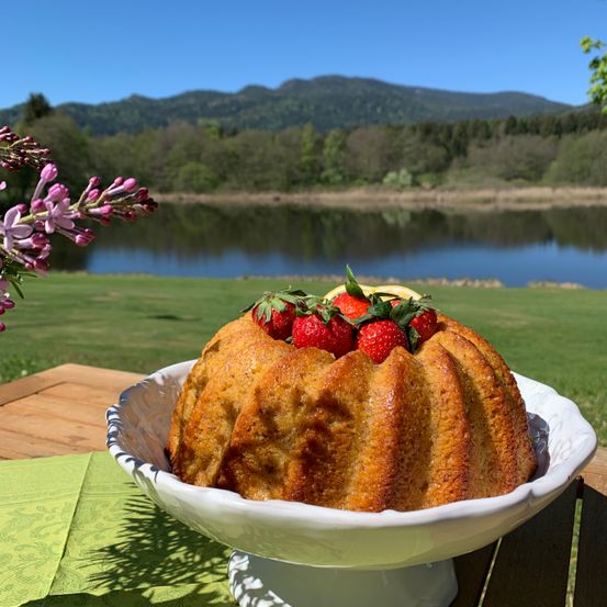 Ein Gugelhupf mit Erdbeeren und Zitronenscheiben auf einem Holztisch am See mit Bergen im Hintergrund.