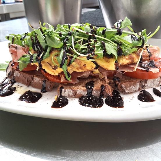 Ein Sandwich mit gebratenen Eiern, Tomaten und Rucola, überzogen mit Balsamico-Glasur auf einem weißen Teller.