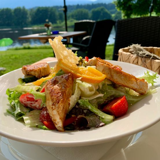 Ein Teller mit gegrilltem Hähnchensalat mit Salat, Tomaten und Orangenscheiben, serviert auf einem Tisch mit Blick auf einen See im Hintergrund.