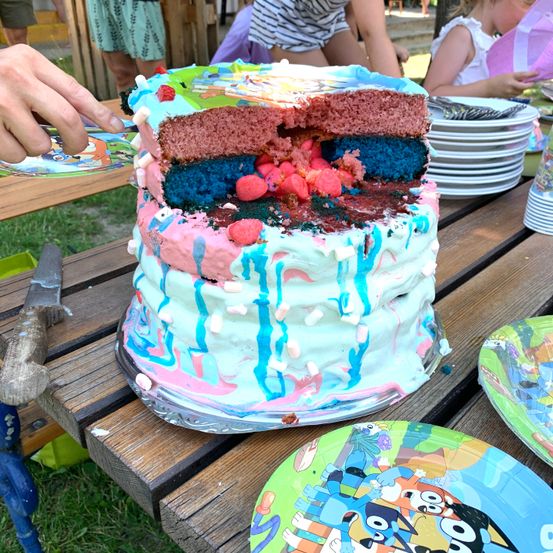 Ein mehrschichtiger Kuchen mit rosa, blauer und grüner Glasur, aufgeschnitten, um seine Schichten zu zeigen, auf einem Picknicktisch mit Tellern und Besteck.