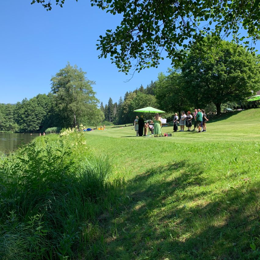 A group of people are gathered on a grassy field near a river, some under an umbrella. Trees and a clear blue sky are in the background.