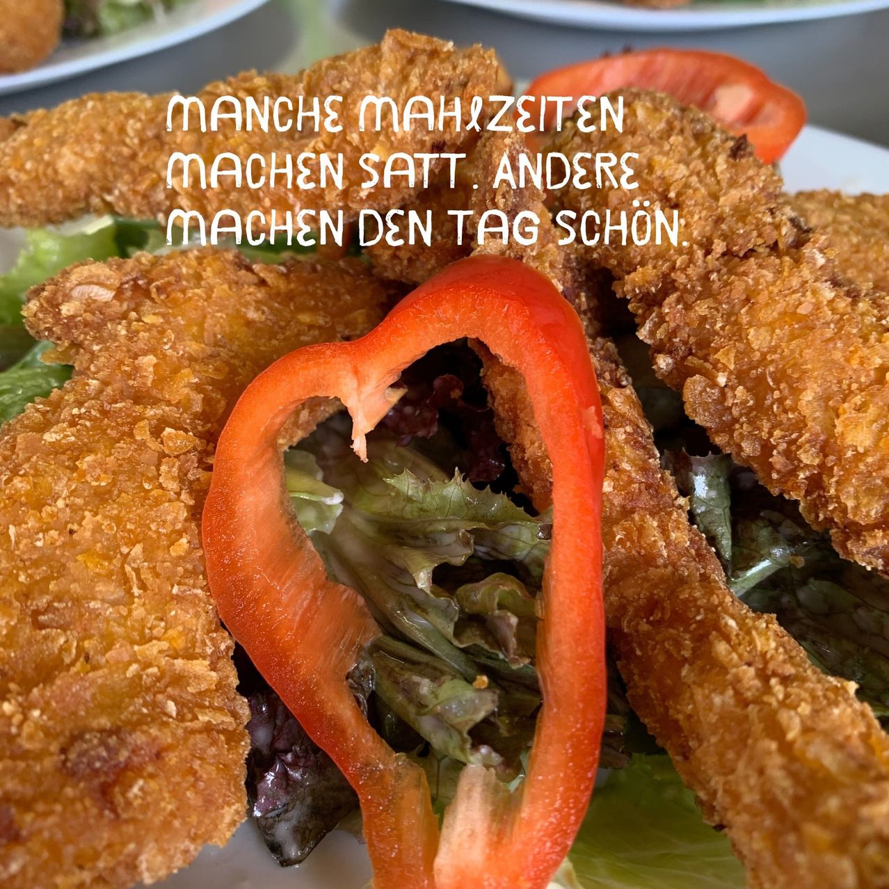 Eine Nahaufnahme eines Tellers mit knusprigen gebratenen Hähnchenstreifen und einer herzförmigen roten Paprika auf einem Bett aus grünem Salat, mit einem deutschen Zitat auf dem Bild.