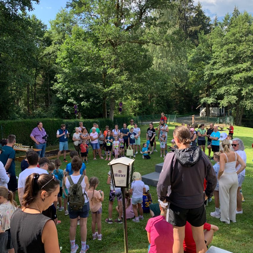 Eine Gruppe von Menschen versammelt sich in einem Park, einige stehen und andere sitzen auf dem Rasen. Eine Person in einer Kapuzenjacke steht in der Nähe eines Schildes. Bäume und ein Haus sind im Hintergrund.