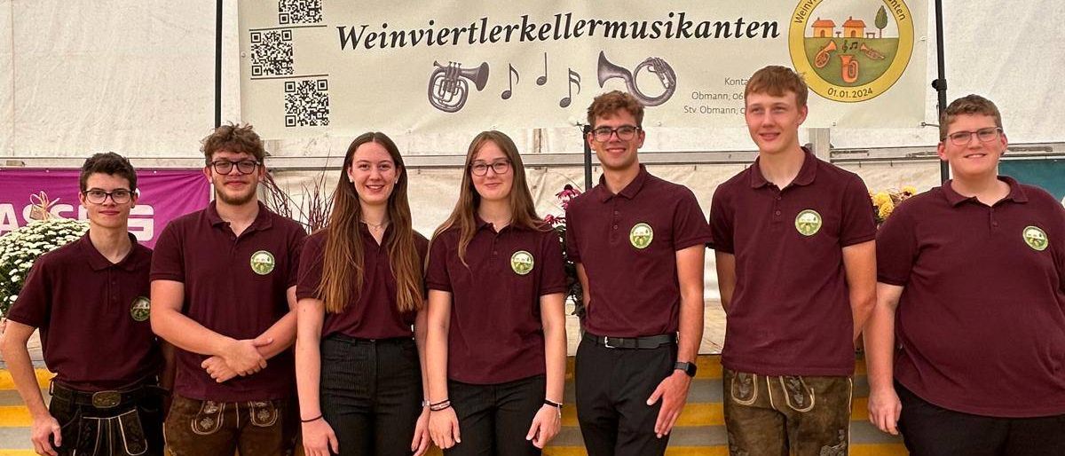 Sechs Personen posieren für ein Foto vor einem Zelt. Sie tragen alle karmesinrote Hemden und schwarze Hosen. Ein Banner mit Text und Musiknoten ist über ihnen aufgehängt.