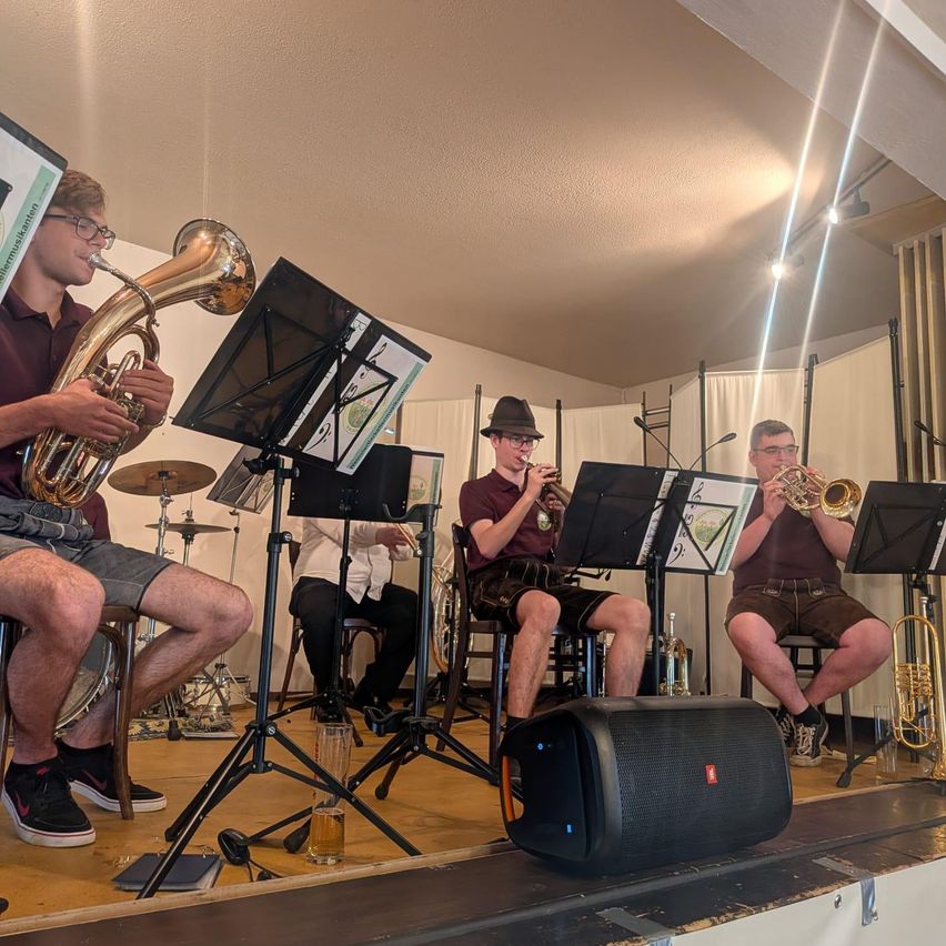 Five musicians are playing brass instruments on stage, with a black speaker in front of them. A person in the front is playing a tuba. They are sitting on chairs in front of music stands.