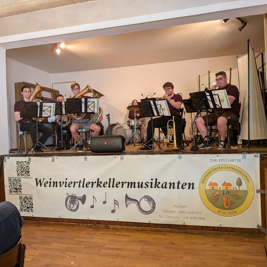 A group of musicians performs on stage with various instruments, including trumpets and drums. A banner with text and music notes is on the stage's front. A speaker and a drum set are on the stage.