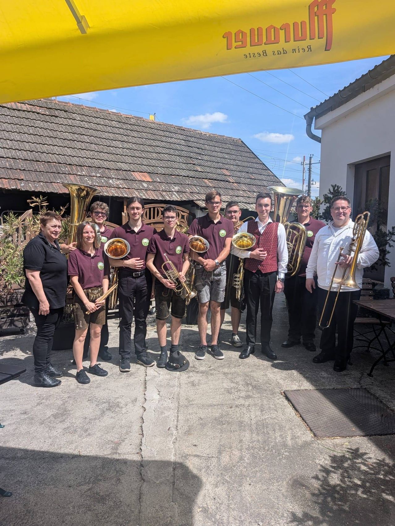 A group of musicians stands outside a house, all holding brass instruments. They are dressed in matching uniforms, suggesting a band. The setting is casual, with a building and plants in the background.