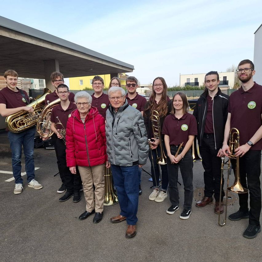 A group of musicians, including a woman and an elderly man, pose for a photo with their instruments in a parking lot.