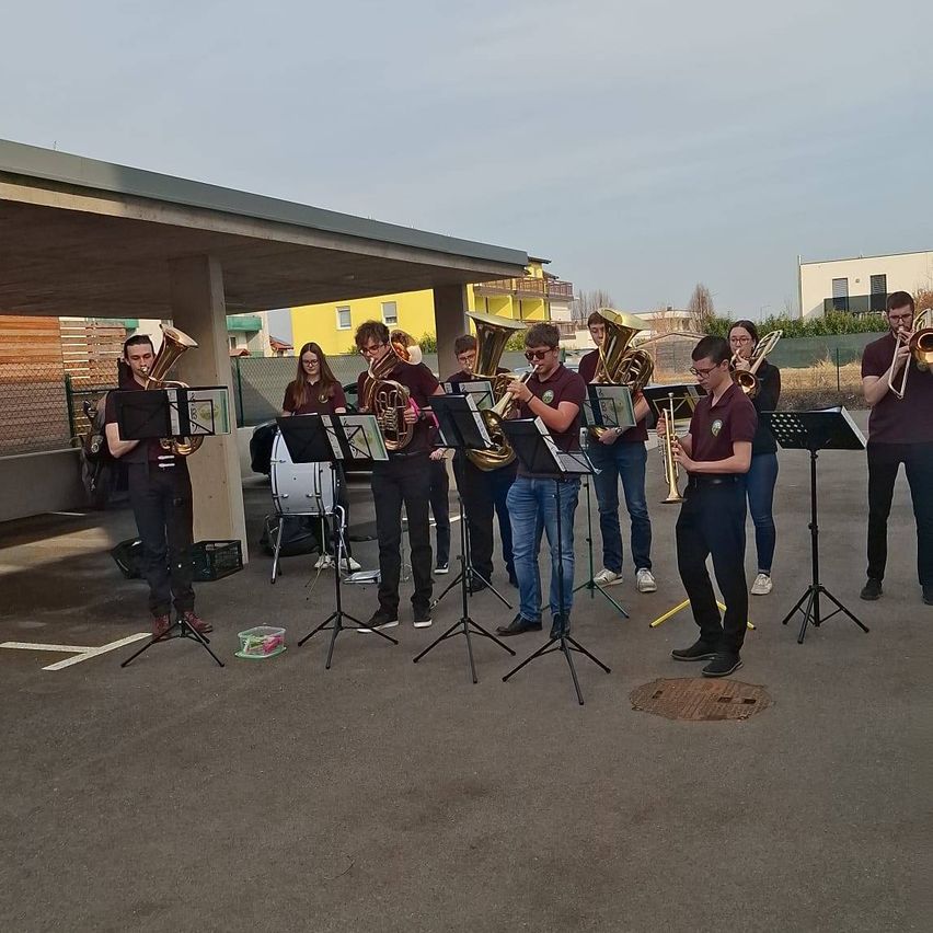A brass band is performing on an outdoor stage with a yellow building in the background.